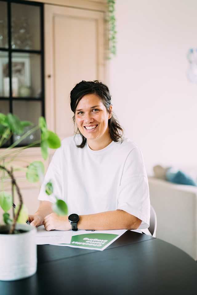 Sofie in een wit shirt glimlacht terwijl ze aan een tafel zit met papieren. Een plant in een pot staat op de voorgrond.