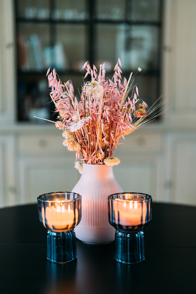 Een witte vaas met gedroogde roze bloemen en twee brandende blauwe kaarsen op een zwarte tafel.